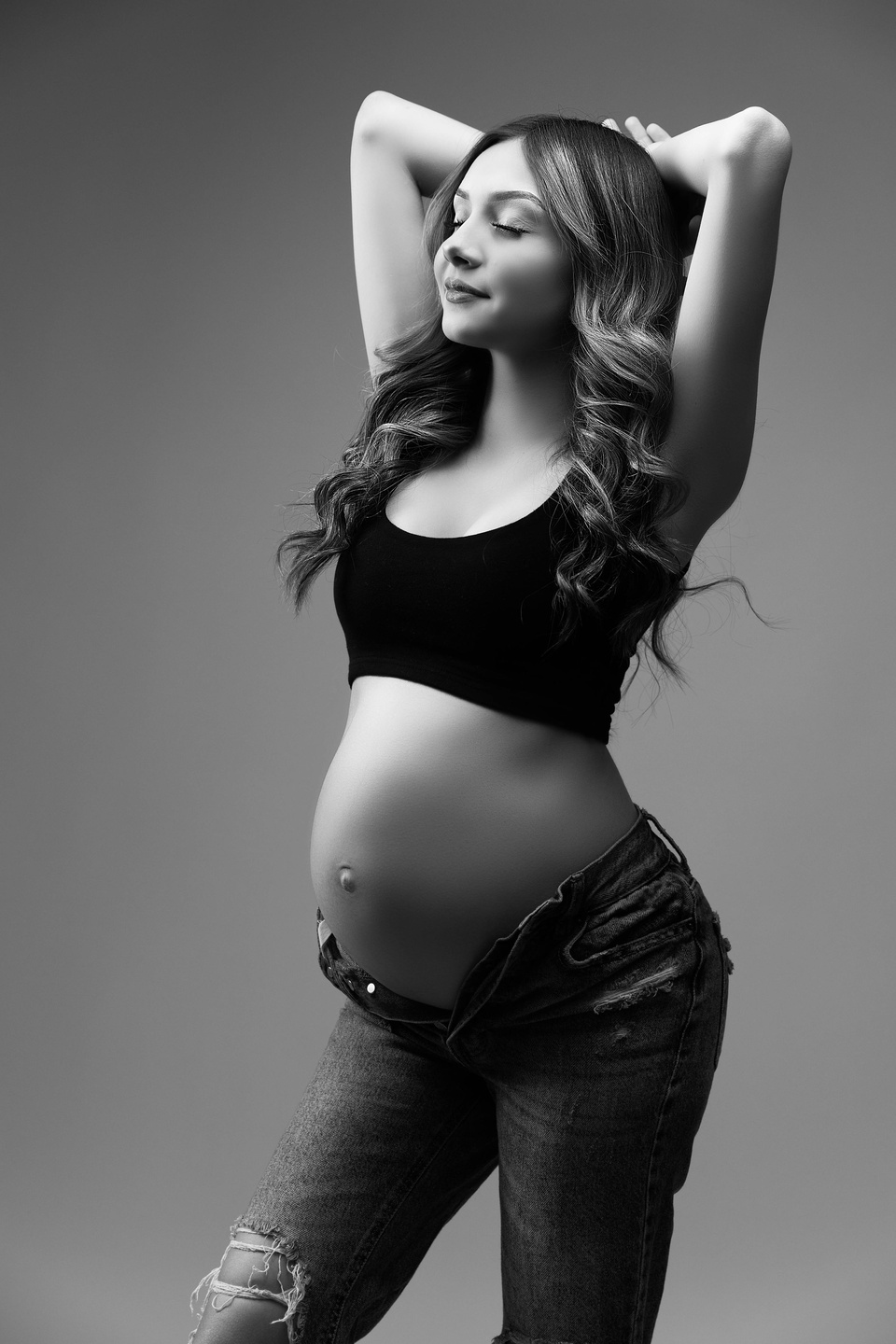 elegant mother with open jeans and a black top exposing her pregnancy belly in a high fashion pose. this portrait was edited in black and white for a timeless luxury look Clavin Klein inspired photography by the best studio photography 