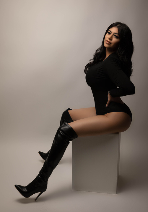 california photographer takes stunning birthday photoshoot of clients wearing black bodysuit and black high boots for a stunning adult birthday photograph with simple and sexy posing long b lack hair and cream backdrop with high end lighting 